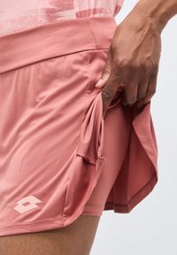 Hånd trækker siden af en støvet pink atletisk skørt og afslører indbyggede shorts med logo på det ydre lag af skørtet.