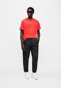 Κόκκινο T-shirt της Nike με μαύρο λογότυπο, συνδυασμένο με μαύρο παντελόνι με κορδόνι στη μέση. Λευκά αθλητικά παπούτσια ολοκληρώνουν το σύνολο, πάνω σε απλό φόντο.