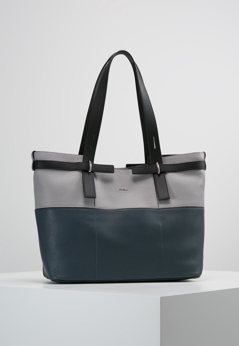 Sac cabas en cuir bicolore gris et bleu foncé avec doubles poignées noires reposant sur un piédestal blanc sur fond uni.