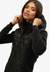Veste zippée noire avec un devant matelassé et capitonné et une capuche. Elle possède des manches longues, deux poches latérales zippées et une texture de tissu lisse.