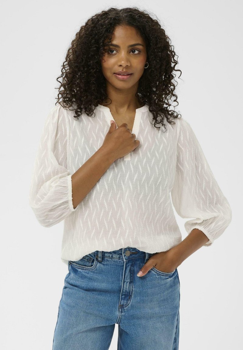 Femme aux cheveux bouclés portant un top blanc texturé et un jean bleu, avec une main touchant sa poitrine et l'autre dans sa poche.