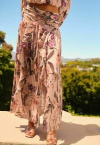 Blumenmuster, leichtes Kleid in zartem Rosa mit dunklen grünen und lila Akzenten. Mit fließendem Stoff und plissiertem Design. Transparente High-Heelsandalen.