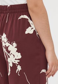 Gros plan sur le bas du torse et le bras d'une personne portant un pantalon bordeaux avec des motifs floraux blancs et une ceinture élastique.
