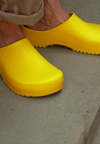 Helle gelbe Clogs mit einer strukturierten Oberfläche, abgerundeten Zehen und dicken Sohlen, die auf einer grauen Betonoberfläche platziert sind.