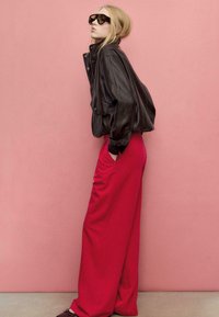 Giacca di pelle nera con collo alto, taglio oversize e dettagli con bottoni abbinata a pantaloni larghi di un rosso acceso. Occhiali da sole scuri completano il look.