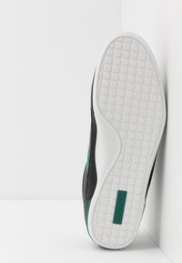 Musta tennari, jossa on teksturoitu pinta, valkoinen kumipohja, jossa on pyöreitä kuvioita, ja vihreä Lacoste-logo kantapäässä.