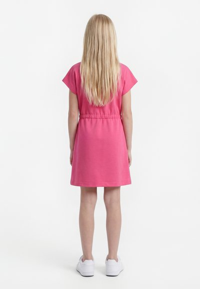 Jeune fille aux longs cheveux blonds portant une robe rose à manches courtes et des baskets blanches, debout, dos à nous sur un fond blanc.