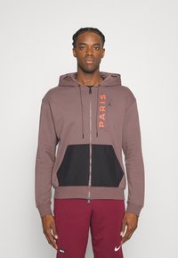 Marsala hoodie med sorte detaljer og kengurulomme, med teksten "PARIS" i orange, lynlås lukning og snoretræk. Blød stoftekstur.