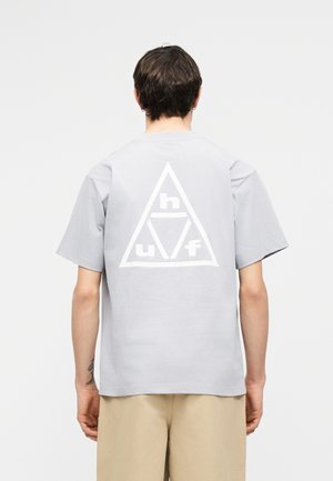 Personne portant une chemise à manches courtes gris clair avec un grand logo triangulaire blanc contenant les lettres « h », « u » et « f » dans le dos, pantalon beige.