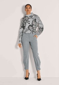 Vrouw in een grijze sweater met bloemenpatroon met lange mouwen, grijze pantalon met omslag en zwarte hakken, staat tegen een effen muur.