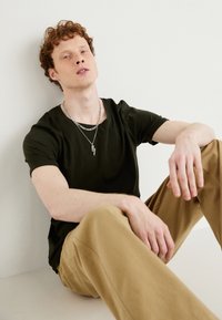 Dark green fitted t-shirt, beige wide-leg pants, layered silver chains, featuring a lightning bolt pendant. Model seated against a light wall.