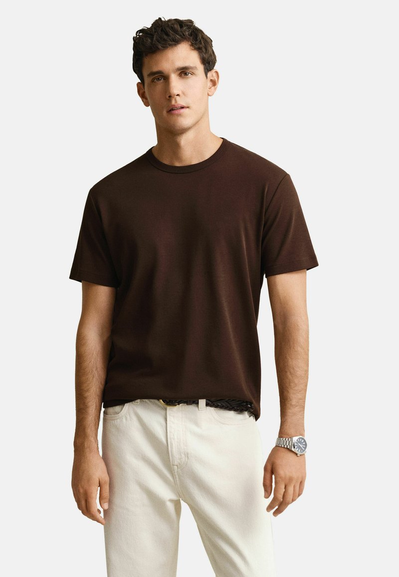 Jeune homme portant un t-shirt à manches courtes marron foncé, un pantalon beige clair, une ceinture tressée et une montre-bracelet argentée, debout devant un fond uni.