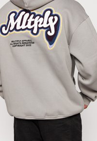Sweat à capuche gris surdimensionné avec texte brodé multicolore au dos, comprenant "Mltply" et des détails de copyright en fine impression.