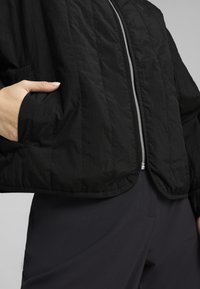 Schwarze gepolsterte Jacke mit einer gesteppte Textur, ausgestattet mit einem silbernen Reißverschluss und seitlichen Taschen. Getragen über dunklen Hosen.