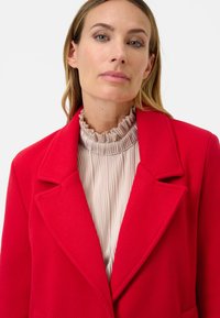 Rode textuur jas met een ingezakte kraag, gecombineerd met een lichtbeige plissékraagtop. Focus op zachte lijnen en contrasterende kleuren.