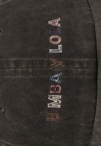 Gorra de pana marrón con texto bordado colorido en diferentes colores de hilo. El diseño incluye un tejido texturizado y detalles de costura distintivos.