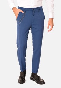 Pantaloni blu su misura con una texture liscia, caratterizzati da una catena a tasca laterale. Indossati con una camicia bianca a maniche lunghe e scarpe nere.