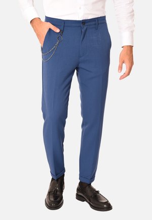 Pantaloni blu su misura con una texture liscia, caratterizzati da una catena a tasca laterale. Indossati con una camicia bianca a maniche lunghe e scarpe nere.