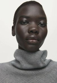 Maglione a dolcevita grigio a coste, texture morbida, design aderente, con collo alto. Pelle liscia e capelli scuri su uno sfondo neutro.