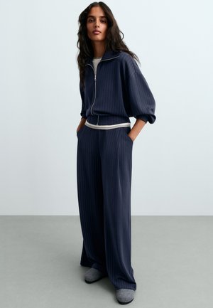 Femme aux longs cheveux foncés portant une veste zippée à rayures bleu marine et un pantalon large assorti, debout les mains dans les poches et des chaussons gris.