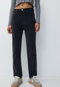 Pantalons en velours côtelé gris foncé à jambes droites, avec des poches avant, une taille haute et une fermeture à bouton en métal, portés avec des baskets de couleur claire.
