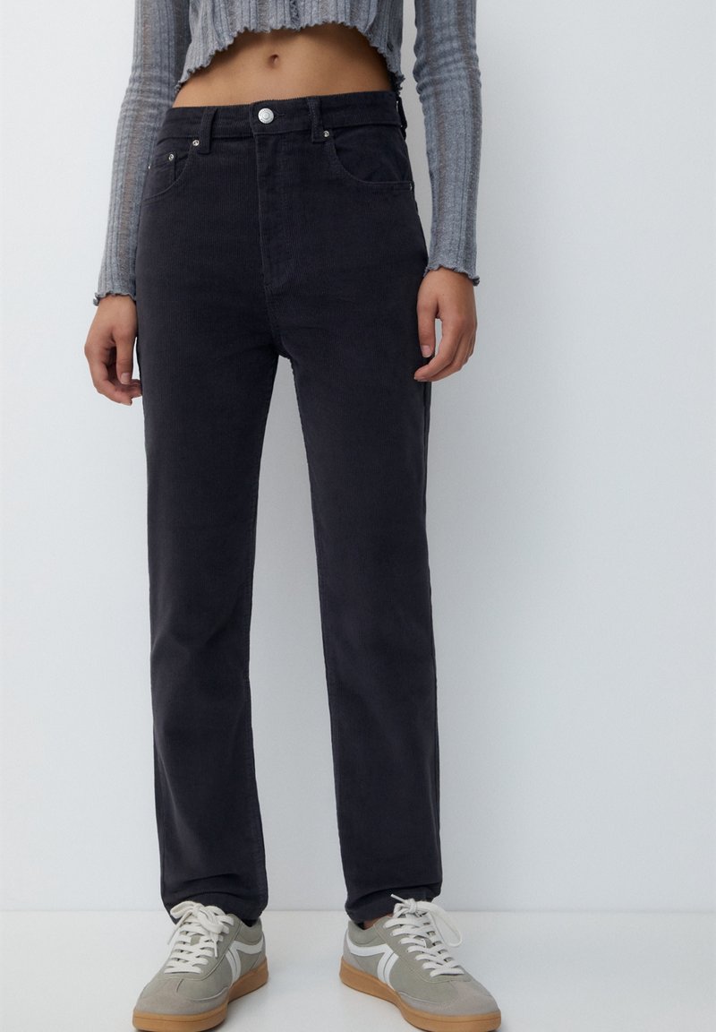 Pantalons en velours côtelé gris foncé à jambes droites, avec des poches avant, une taille haute et une fermeture à bouton en métal, portés avec des baskets de couleur claire.
