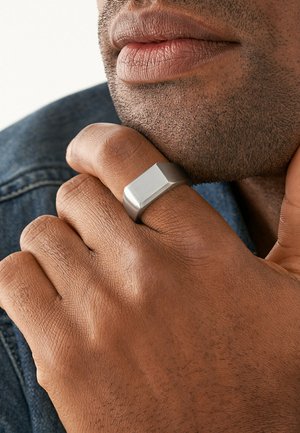 Anello in metallo con un design geometrico piatto. Finitura opaca in grigio argento. Indossato su una mano con pelle texturizzata che riposa vicino al mento.
