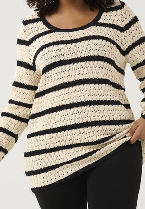 Woman wearing a cream and black striped knit sweater with a scoop neck, holding the hem, paired with black pants.