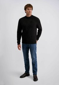 Zwarte gebreide trui met een hoge ronde hals, gecombineerd met een blauwe spijkerbroek. De outfit heeft een ontspannen pasvorm en ribgebreide manchetten, aangevuld met donkere schoenen.