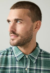 Green plaid flannel shirt with a button-down collar, featuring white and navy stripes, made of textured fabric. Thin buttons visible.