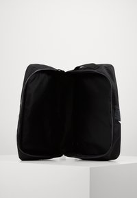 Svart Nike mjuk sidor resväska öppen och tom, placerad på en vit yta med en plain ljusgrå bakgrund.