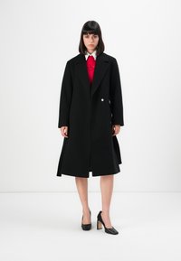 Donna in piedi che indossa un lungo cappotto nero sopra un maglione rosso e una camicia bianca, abbinati a scarpe con tacco alto nere, su uno sfondo semplice.