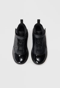 Baskets de sport noires avec une finition en vernis brillant, un dessus en matière texturée mate, des lacets élastiques et une languette. Présente un logo sur la sangle.