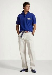 Jeune homme portant un polo bleu, un pantalon blanc, une ceinture rayée et des baskets bleues, se tenant les mains dans les poches, regardant sur le côté.