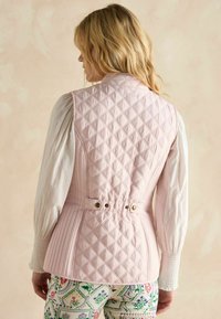 Rosa steppierte Weste mit einer taillierten Rückengestaltung, ausgestattet mit goldfarbigen Hardware-Elementen und einem strukturierten Stoff. Darunter ein langärmliges weißes Hemd.