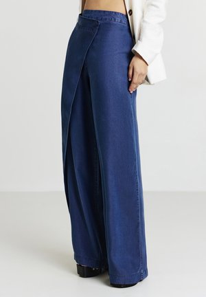 Person in high-waisted, wide-leg blauen Hosen mit überlappendem Vorderteil, weißem Langarmoberteil und schwarzen Plateauschuhen.