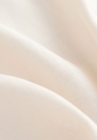 Měkká bílá lněná látka s viditelnou tkanou strukturou a jemnými záhyby vytvářejícími hladké, plynule tekoucí vlny.