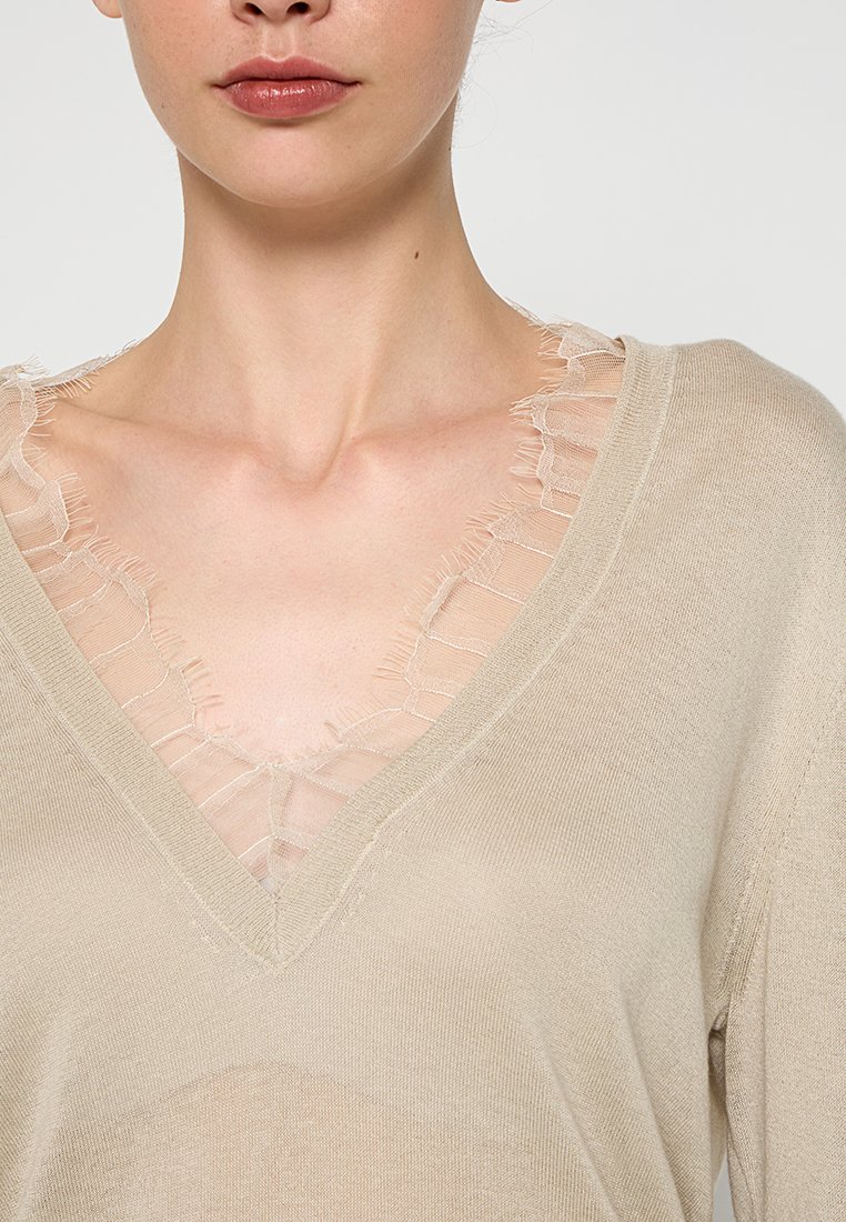 Beige gebreide top met een V-hals en doorschijnende rafelige kanten rand. Heeft een ontspannen pasvorm en een zachte textuur. Geen opmerkelijke patronen of hardware zichtbaar.