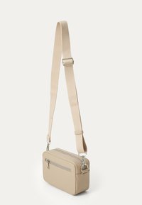 Sac bandoulière beige en matériau lisse. Doté d'un haut zippé, d'une poche avant et d'une bandoulière réglable avec des accents en métal argenté.