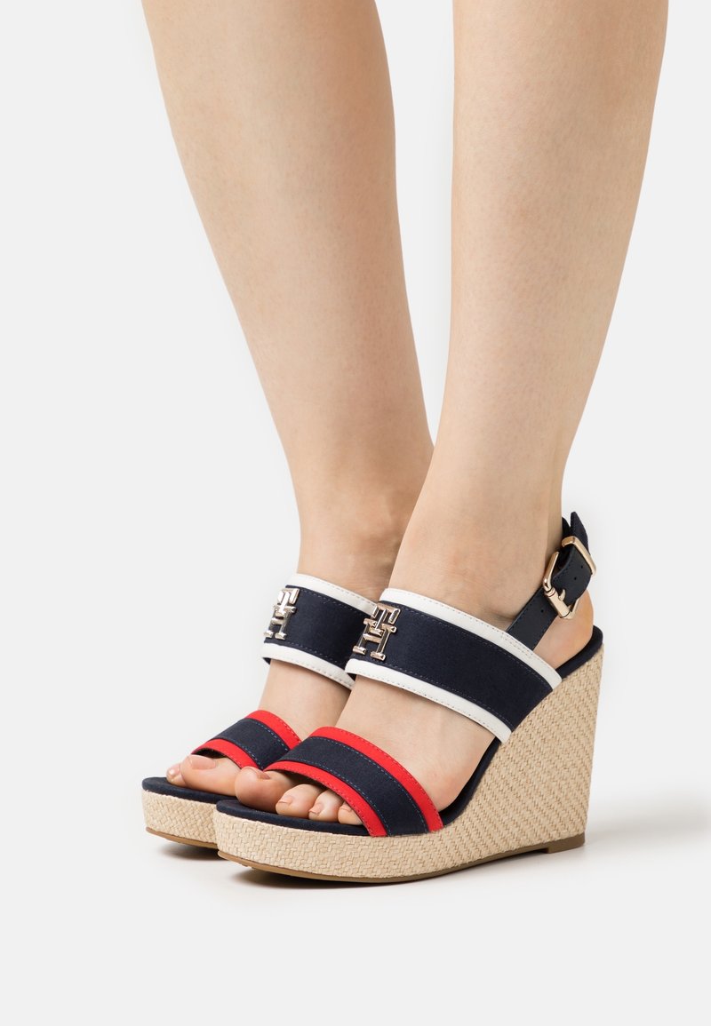 Pieds portant des sandales compensées à bout ouvert avec des lanières bleu marine, blanc et rouge et un talon beige texturé sur un fond uni.