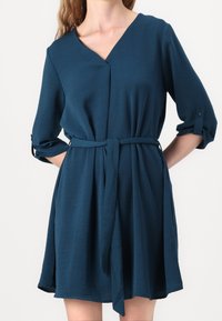 Robe col V couleur sarcelle en tissu texturé, avec manches trois-quarts, ceinture à la taille et ourlet évasé.