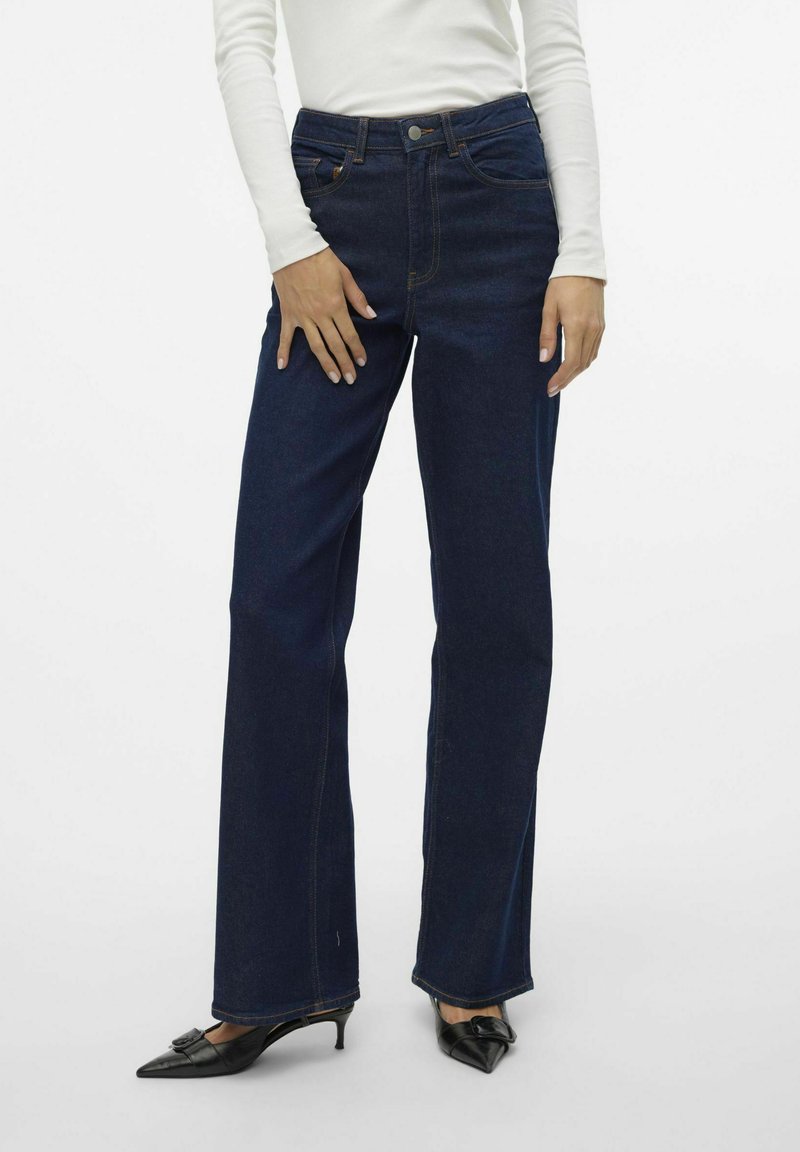 Mörkblå högmidjade jeans i en utsvängd design, med framfickor och kontrasterande sömmar, tillsammans med svarta pumps med spetsig tå.