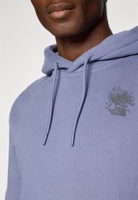 Nærbillede af en person iført en lysepurpur hoodie med snore og et mørkt abstrakt logo på brystet.