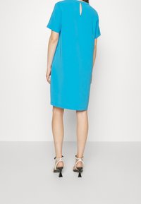Tyrkysové šaty s krátkým rukávem a detaily ve tvaru klíčové díry na zádech, vyrobené z hladkého materiálu. Nosí se s bílými a černými sandály na podpatku.