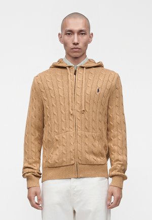 Homme portant un sweat à capuche zippé beige en maille torsadée par-dessus une chemise rayée à col et un pantalon blanc sur un fond blanc uni.