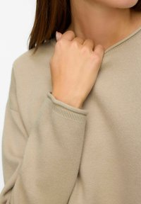 Maglione beige con lavorazione a maglia testurizzata, scollatura rotonda e polsini a costine, parzialmente mostrato con una mano che afferra il tessuto vicino al collo.