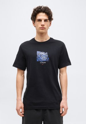 Junger Mann mit lockigem Haar, der ein schwarzes T-Shirt trägt, auf dem ein blaues Paris Saint-Germain-Logo und der Schriftzug "ici c'est paris." abgebildet sind.