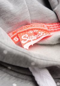 Tessuto felpato grigio con piccoli pois bianchi, completo di un'etichetta arancione con la scritta "SuperDry" e "Vintage" in bianco.