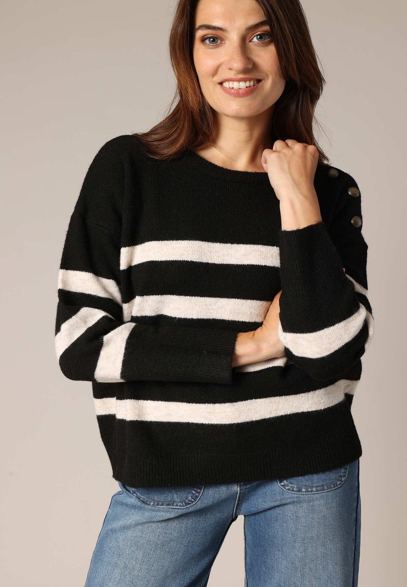 Pull à rayures noir et blanc, texture tricotée, col rond, avec des boutons décoratifs sur l'épaule, associé à un jean bleu.