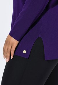 Close-up van een persoon die een paarse lange mouw top met een zijsplit en een ronde gouden logo draagt, in combinatie met zwarte broek, hand rustend op de dij.